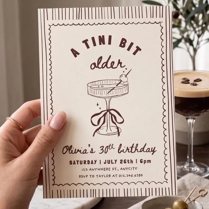 A Tini Bit Older birthday invitation on cream card in cocoa ink, with a hand-drawn espresso martini coupe tied with a bow inside a stamp-edge border