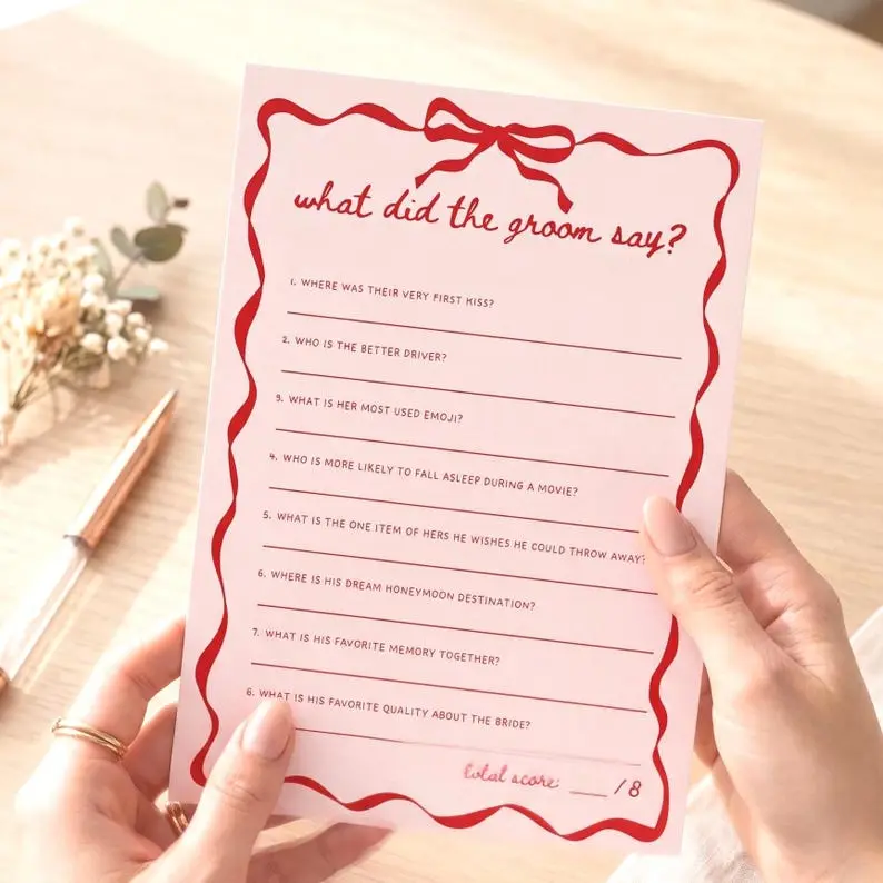 What Did The Groom Say game card in blush pink with a red wavy ribbon border and bow, listing eight questions on ruled lines with a score box at the foot